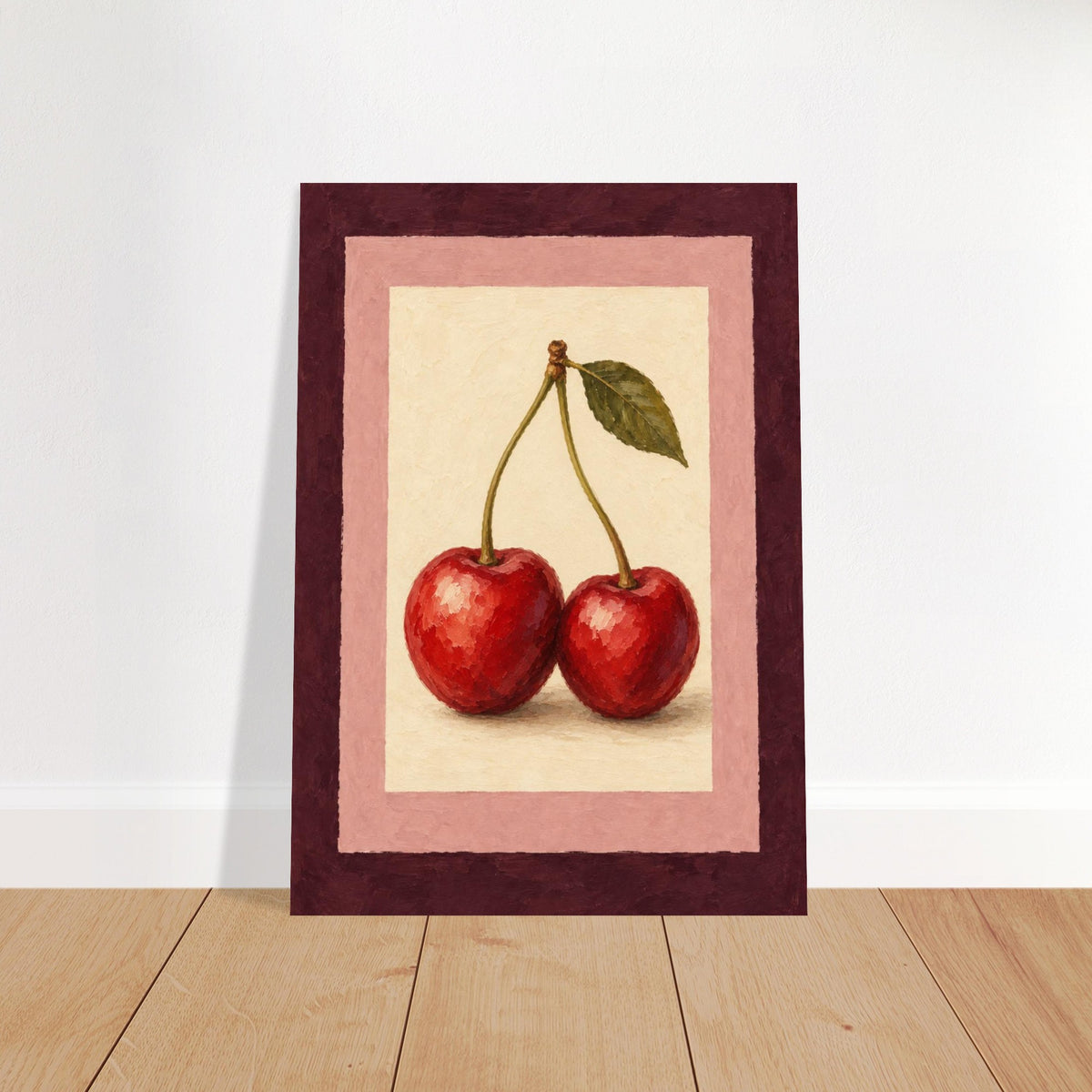 A framed art print featuring a still life painting of two red cherries with green leaves, set against a cream background with pink and dark burgundy borders, leaning against a white wall on a light wood floor.