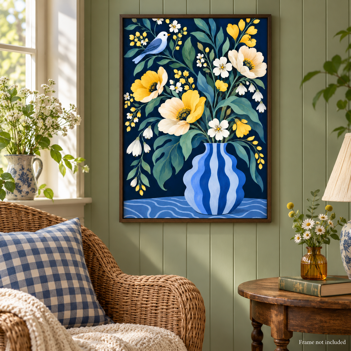 A framed floral art print hangs on a green paneled wall above a wicker armchair in a styled living room. The artwork depicts a blue striped vase filled with yellow and white flowers, green leaves, and a small blue bird on a dark blue background.