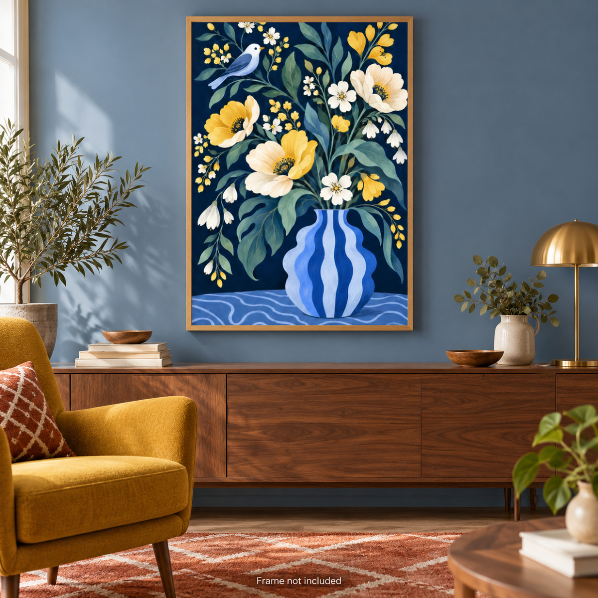 A framed floral art print hangs on a blue wall above a wooden console table in a styled living room. The artwork shows a blue striped vase with yellow and white flowers, green leaves, and a small blue bird.