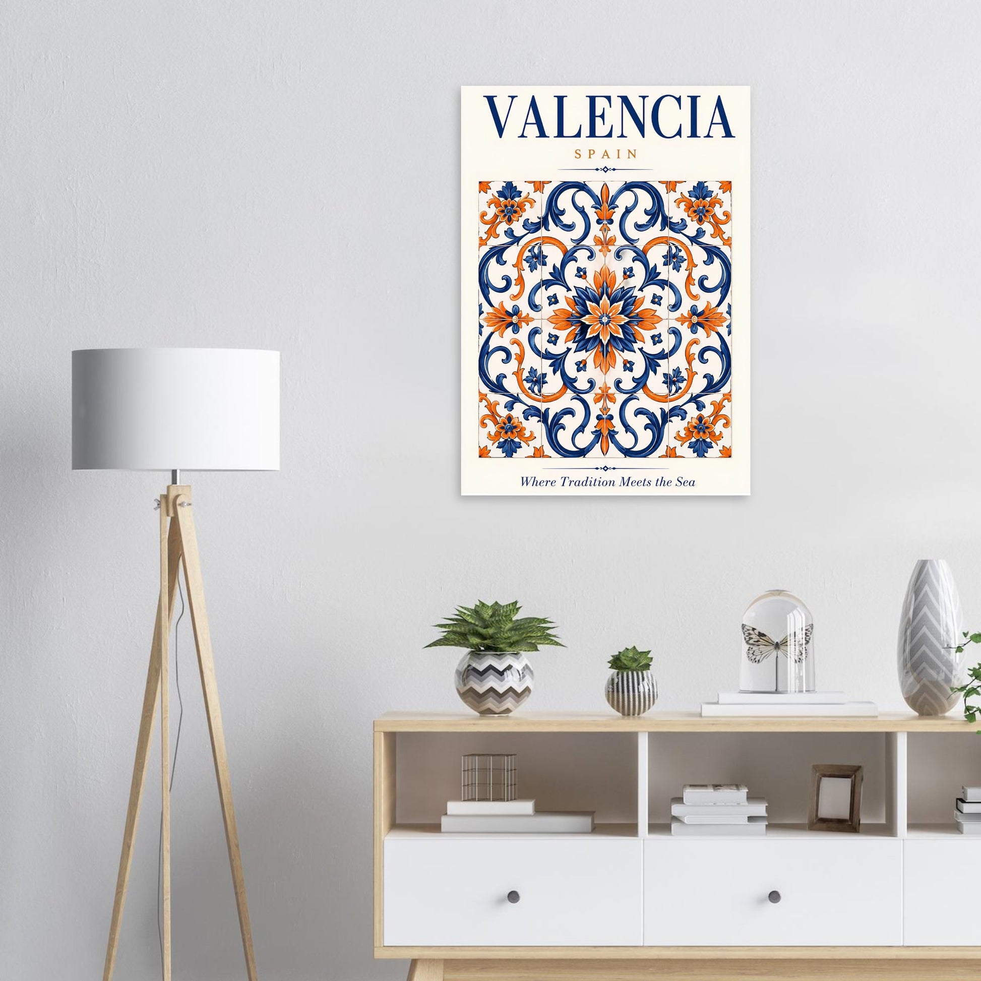 A framed wall art print featuring a decorative Valencia, Spain tile pattern in navy blue, orange, and cream, displayed above a light wood and white cabinet in a styled room with a floor lamp and small plants.