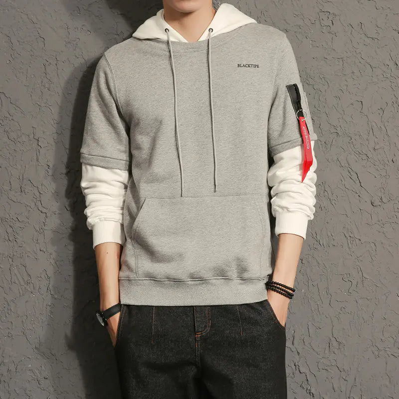 Person wearing a gray hoodie with white sleeves and red accents against a gray textured wall.