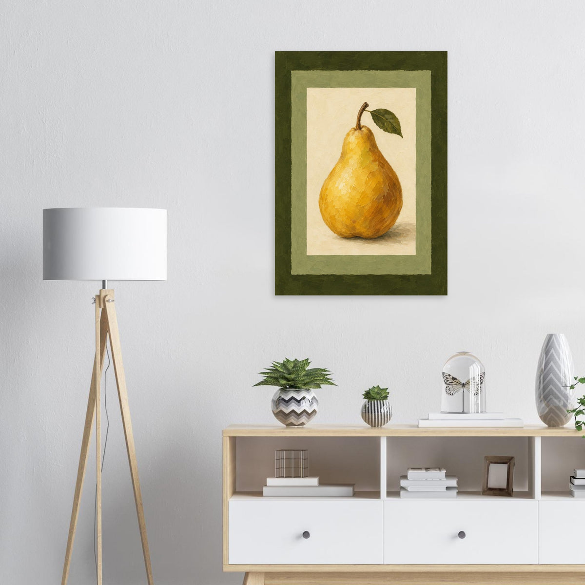 A framed vertical art print of a yellow pear with a green leaf, displayed on a white wall above a light wood console cabinet with decorative plants and a tripod floor lamp.