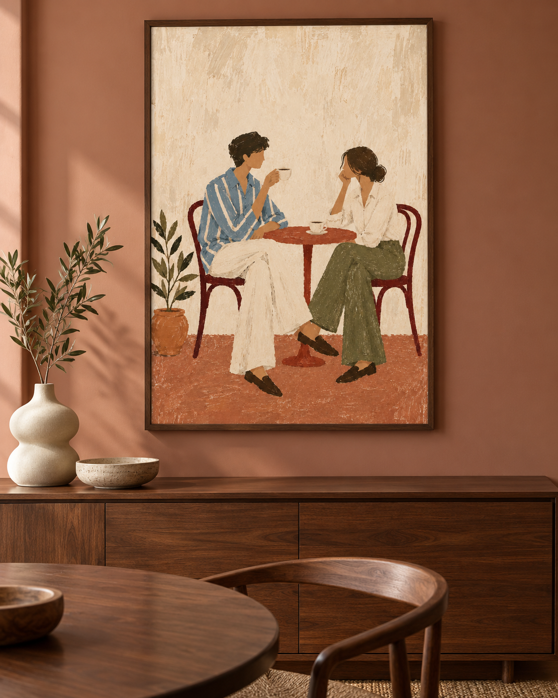 A framed art print depicting a couple sitting at a small table, displayed on a terracotta-colored wall above a wooden sideboard in a styled room.