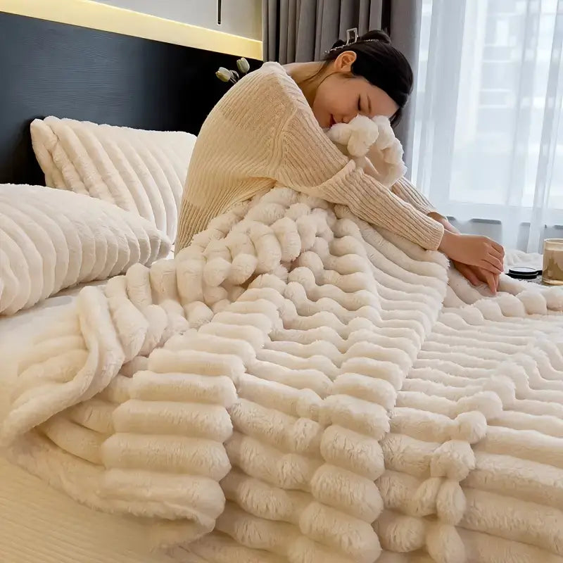 Woman wrapped in a luxurious cream-colored faux rabbit fur blanket, sitting on a neatly made bed in a softly lit bedroom.