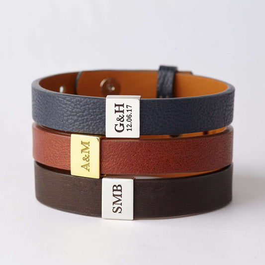 Three stacked leather bracelets in black, brown, and navy with engraved silver and gold charms showing initials and dates – personalized gift for men or couples.