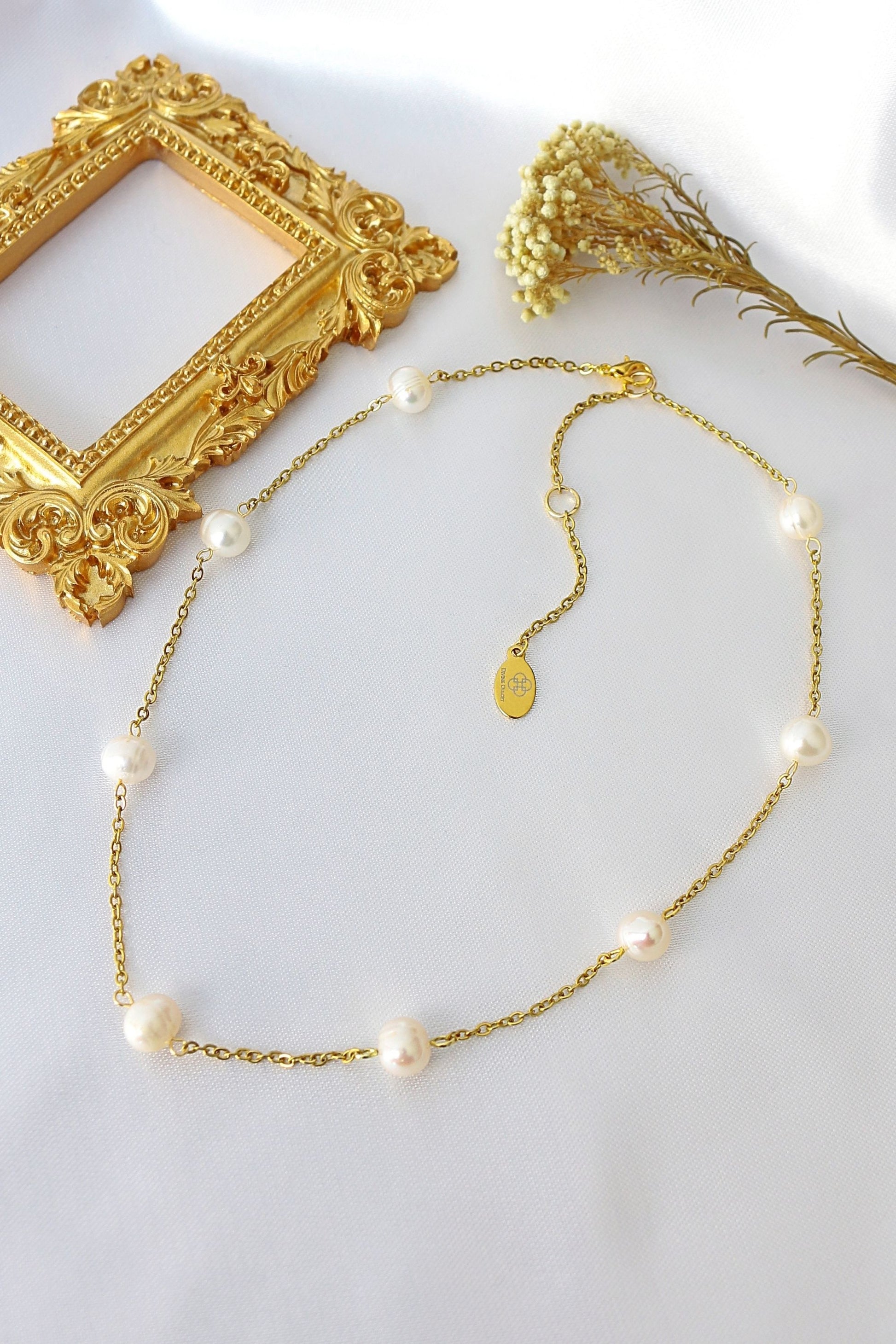Gold necklace with pearls on a white surface with a decorative gold frame and dried flowers.