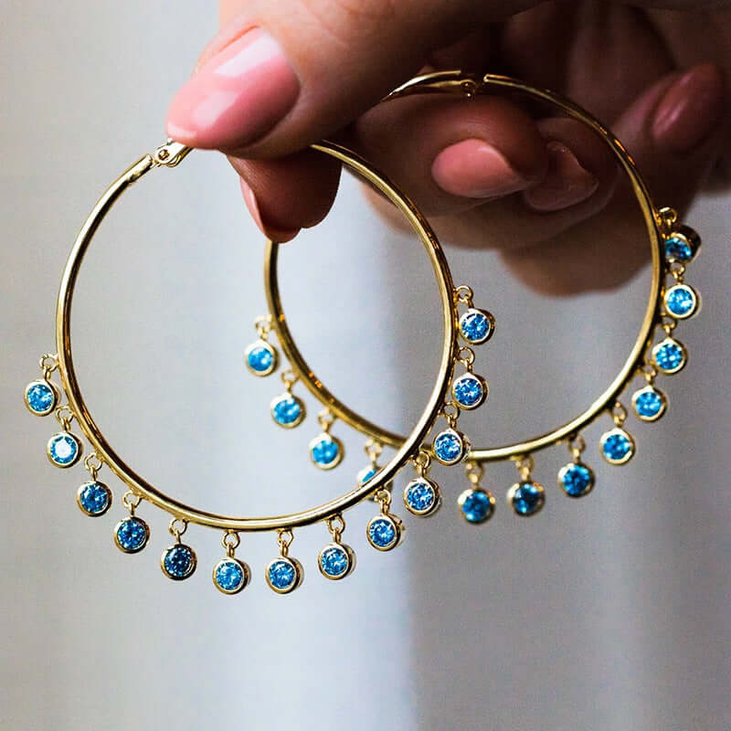 Gold bohemian style hoop earrings with dangling blue gemstone accents held by hand in front of a neutral background