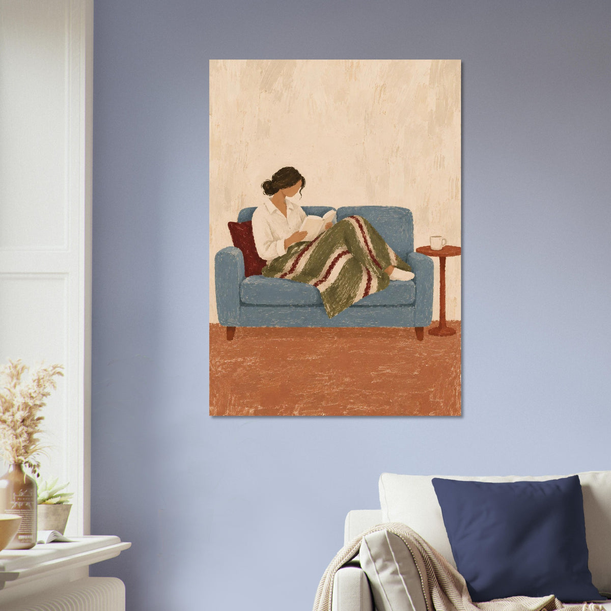A vertical canvas wall art print depicting a woman reading on a blue couch, hung on a light blue-gray wall above a white sofa with a navy pillow.