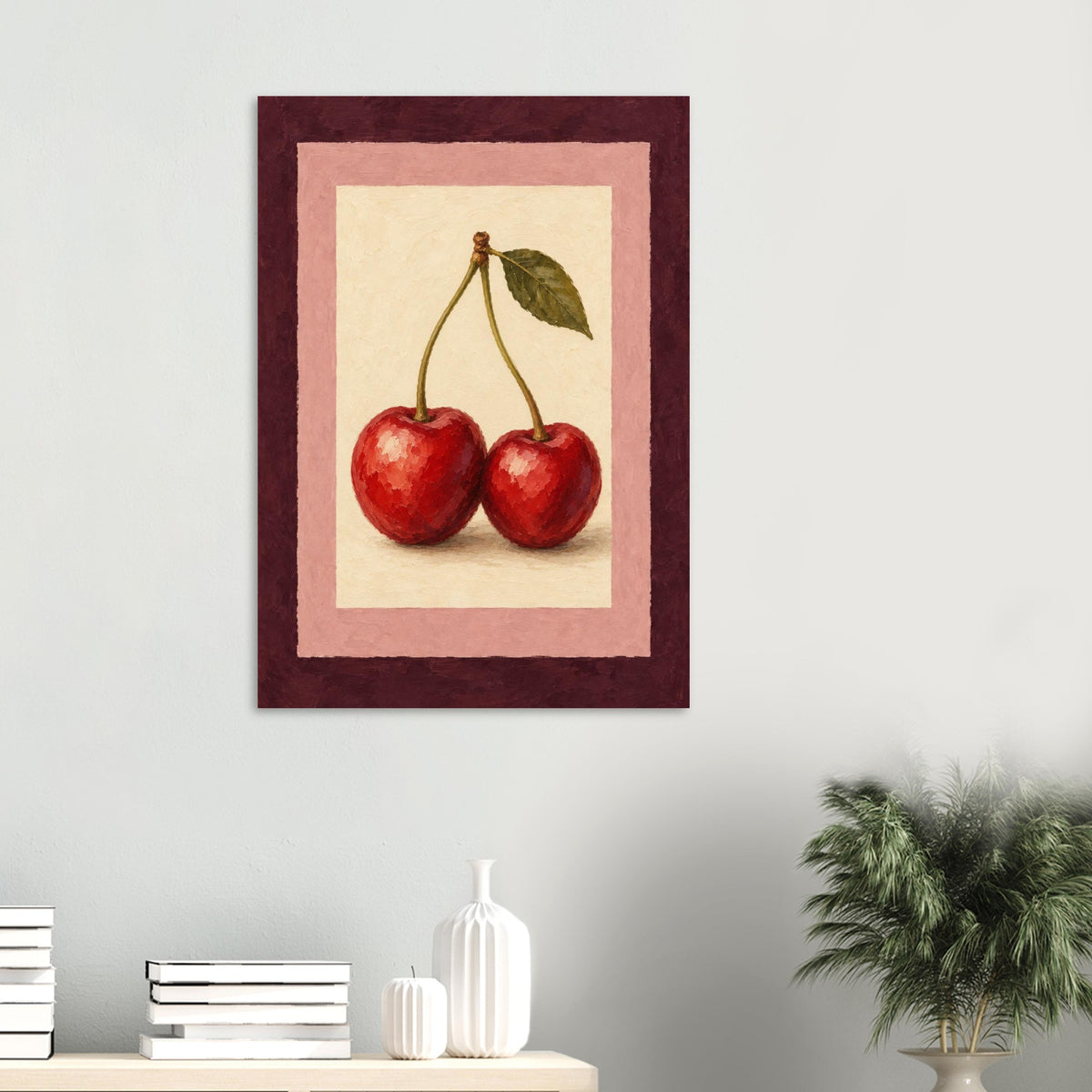 A framed wall art print featuring two red cherries with a green leaf, displayed on a light gray wall above a shelf with books and decorative vases, with a potted plant on the right.
