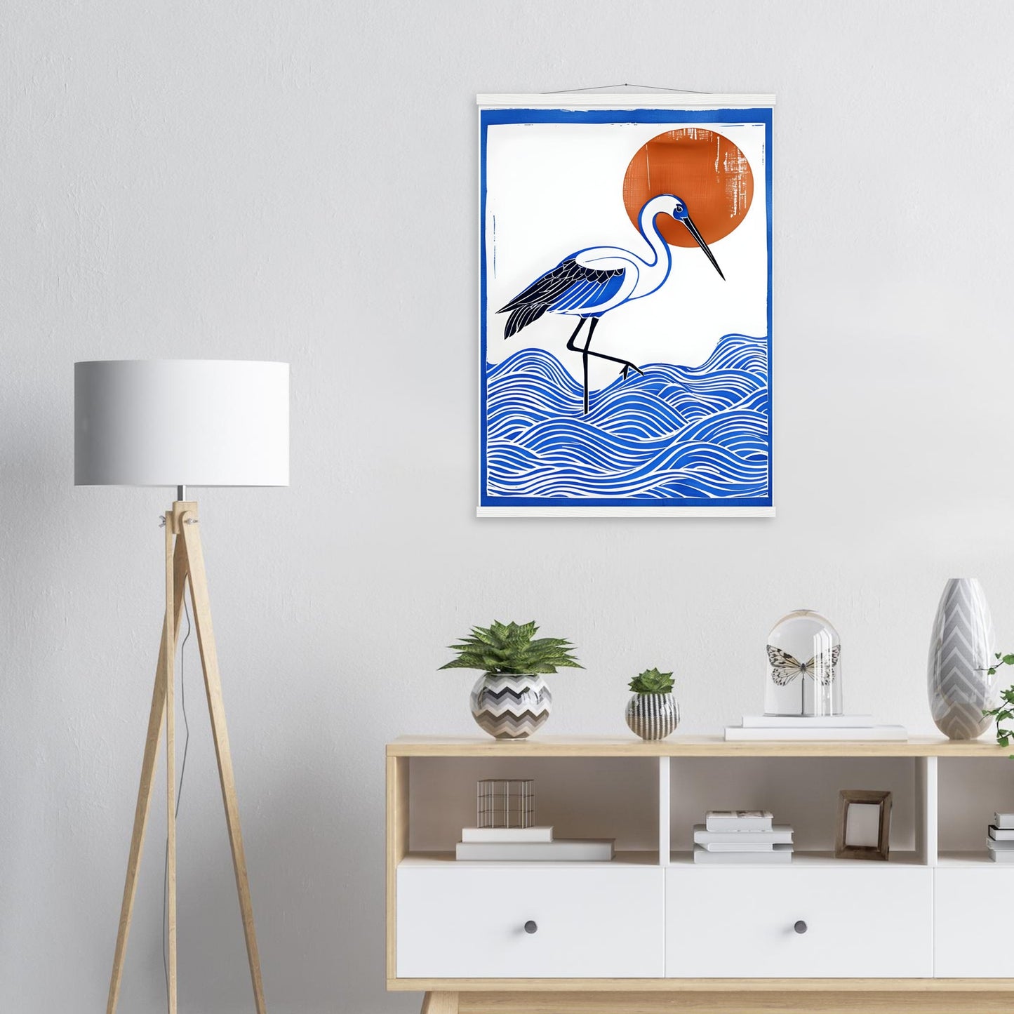 Framed artwork of a crane bird on waves with a red sun above, displayed in a room with a lamp and shelves.