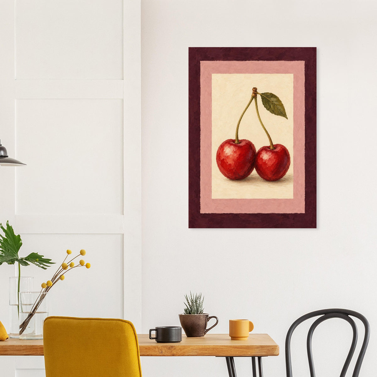 A framed wall art print depicting two red cherries with a green leaf, hung on a white wall above a wooden dining table with chairs and mugs.