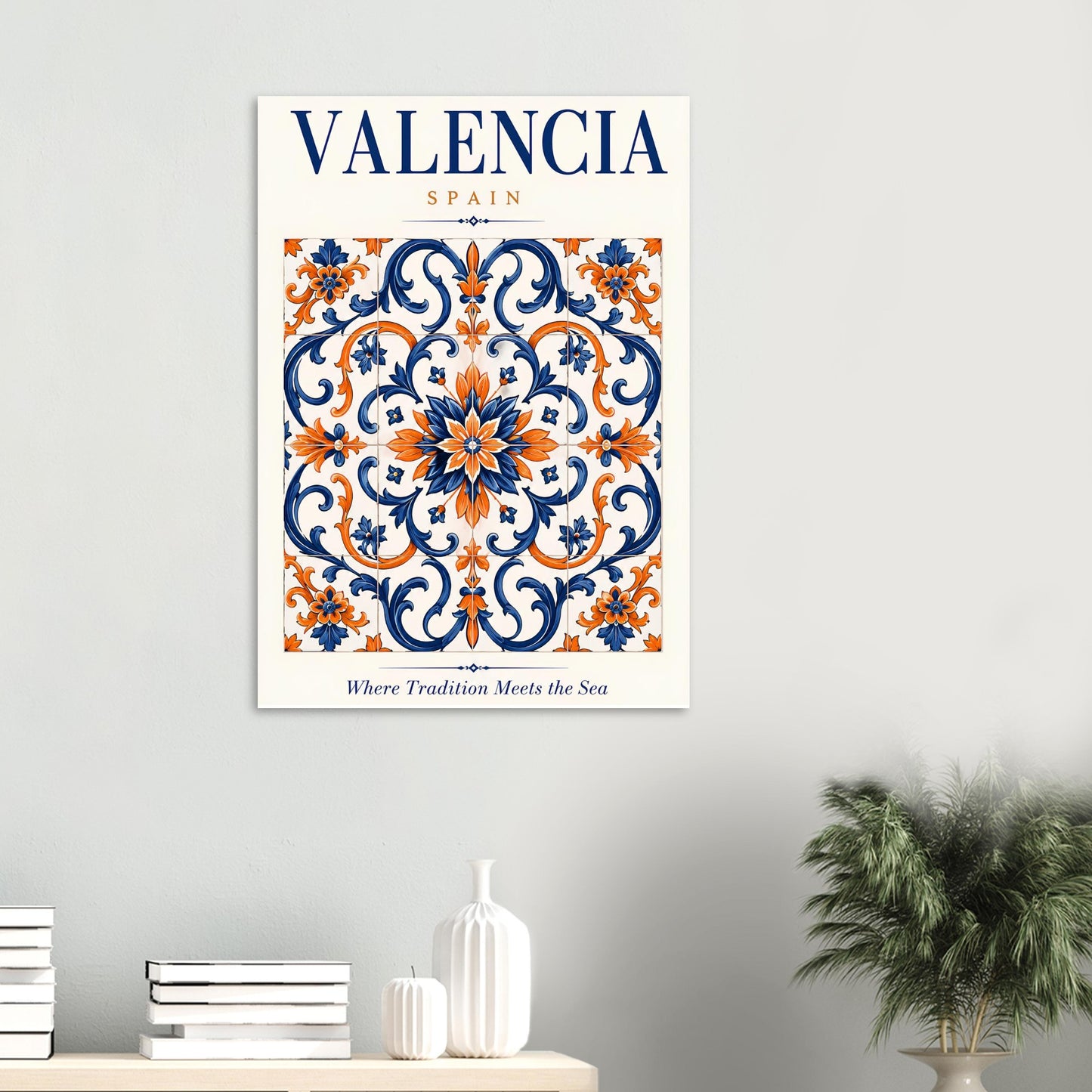 A framed wall art print featuring a decorative Valencia, Spain tile pattern in navy blue, orange, and cream, hung on a light gray wall above a console table with books and a plant.