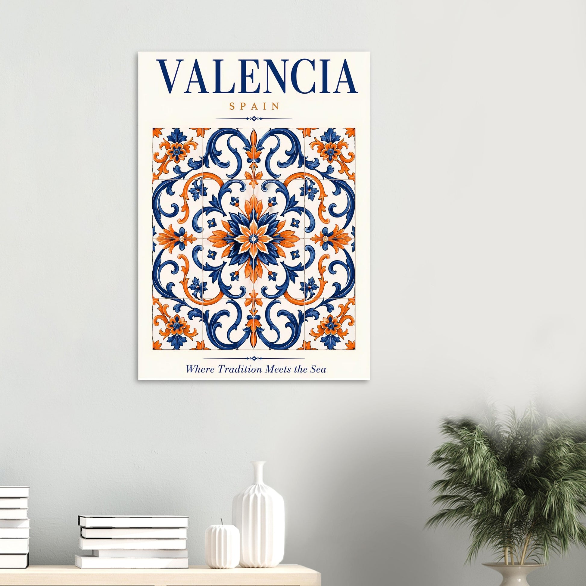 A framed wall art print featuring a decorative Valencia, Spain tile pattern in navy blue, orange, and cream, hung on a light gray wall above a console table with books and a plant.