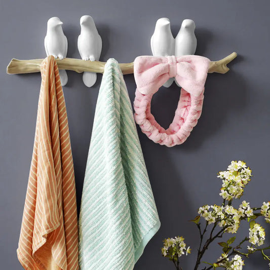 Decorative wall hook with bird designs holding towels and a pink ring against a gray background.