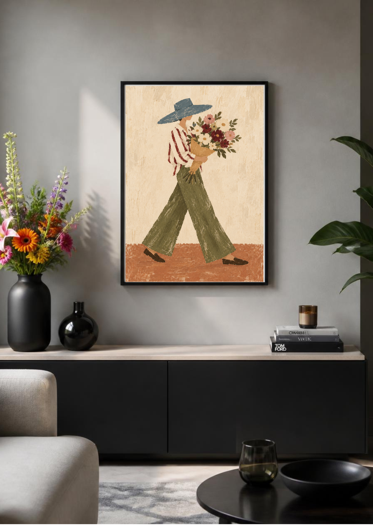 A framed art print depicting a woman in a wide-brimmed hat and green pants carrying a bouquet of flowers, styled on a gray wall above a black console table in a modern living room setting.