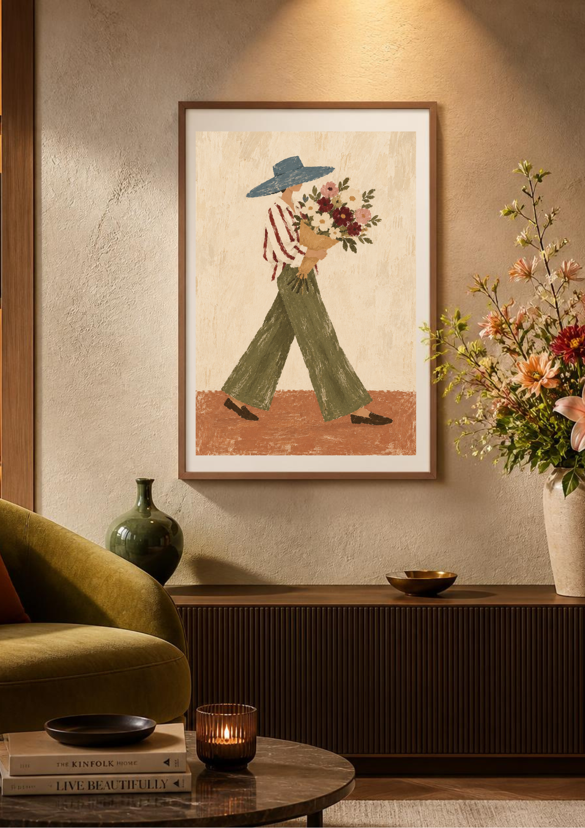 A framed art print featuring a stylized illustration of a woman in a blue hat and green pants carrying flowers, displayed on a textured beige wall in a cozy living room setting with a green armchair, sideboard, and floral arrangements.