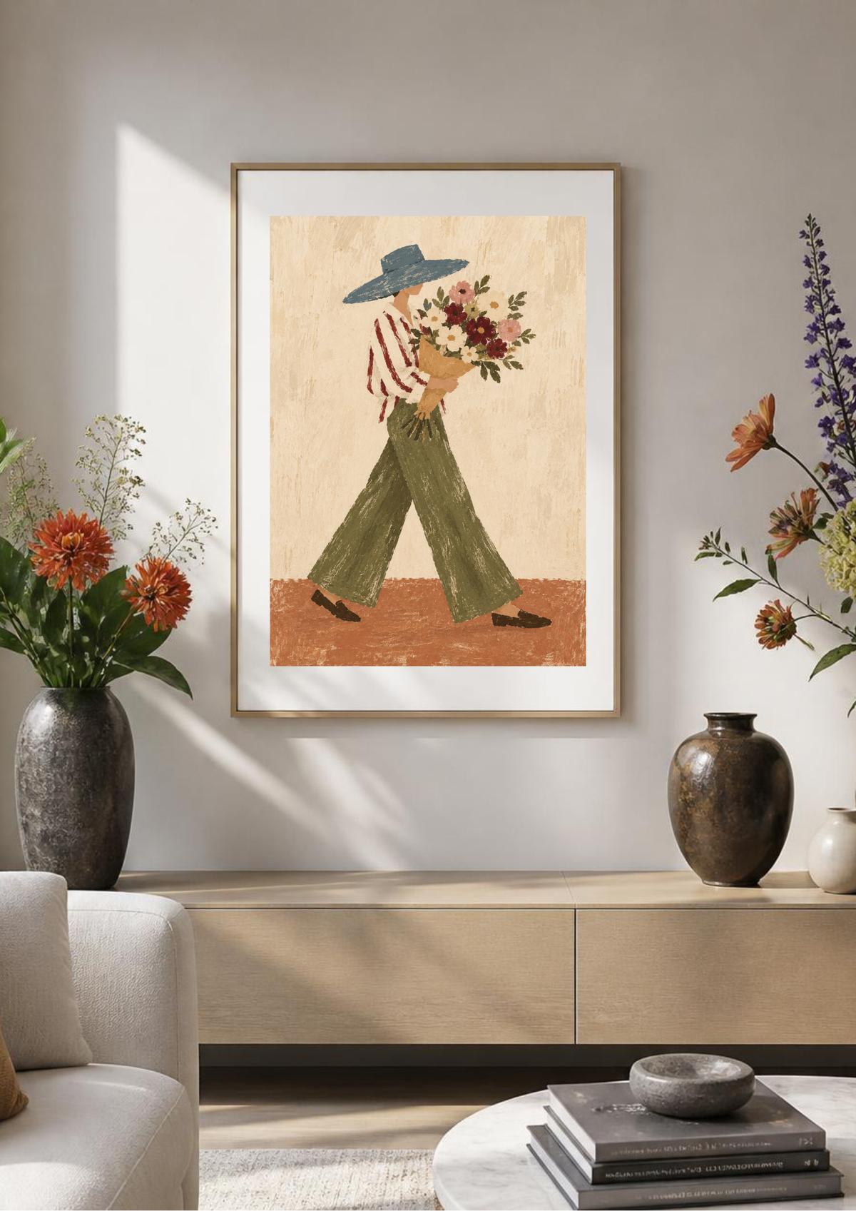 A framed art print featuring a whimsical illustration of a person in a blue hat and green pants carrying a bouquet of flowers, styled in a modern living room setting above a light wood console table.