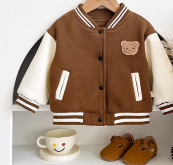 Children's brown jacket with bear emblem on a hanger, next to a smiley face mug and brown shoes.