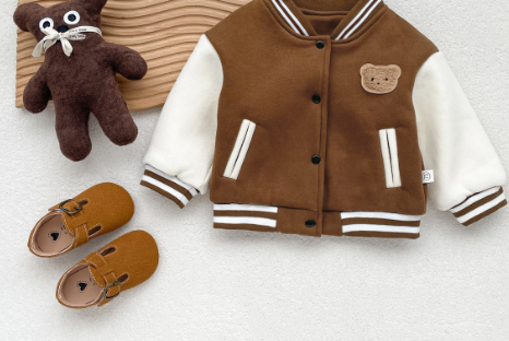Children's brown jacket with white sleeves, teddy bear toy, and brown shoes on a light gray background