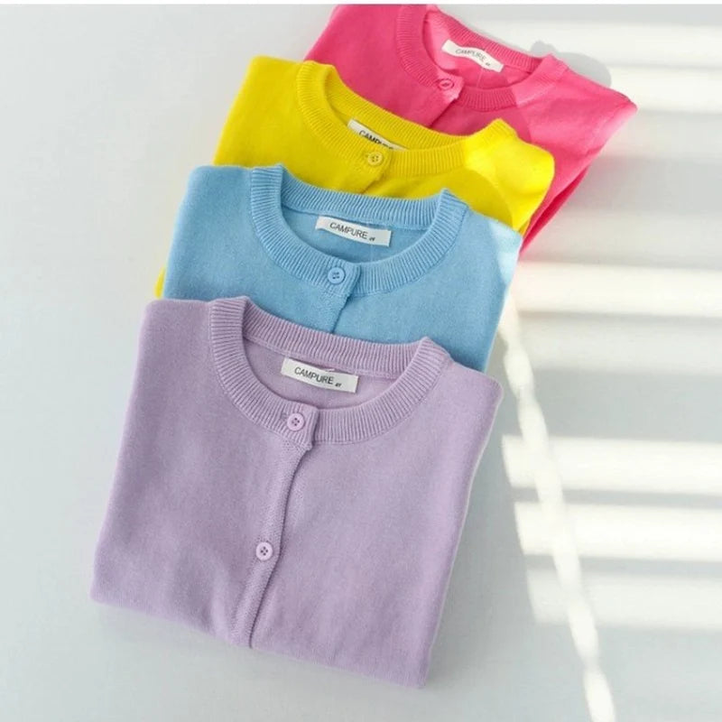 Four folded children's cardigans in purple, blue, yellow, and pink on a light gray background.