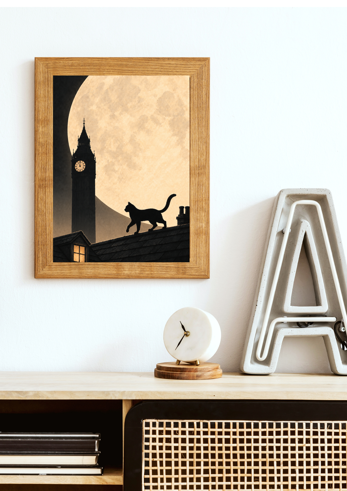 A framed art print featuring a black cat silhouette walking along a roof in front of a large full moon and big ben, displayed on a light wall above a wooden console table with a small clock and a decorative letter A.
