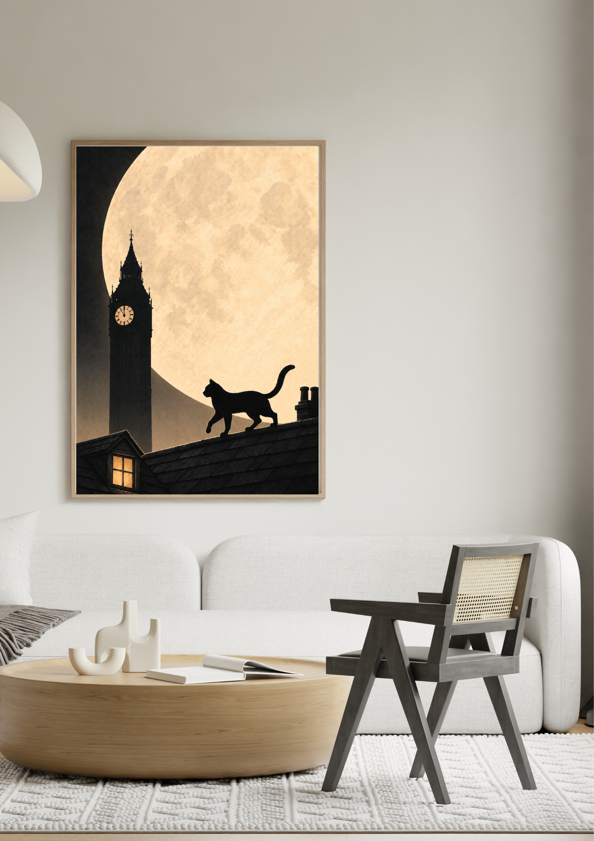A framed vertical art print featuring a black silhouette of a cat walking on a rooftop in front of a large cream-colored full moon and big ben, displayed on a light gray wall above a modern white sofa and wooden coffee table.