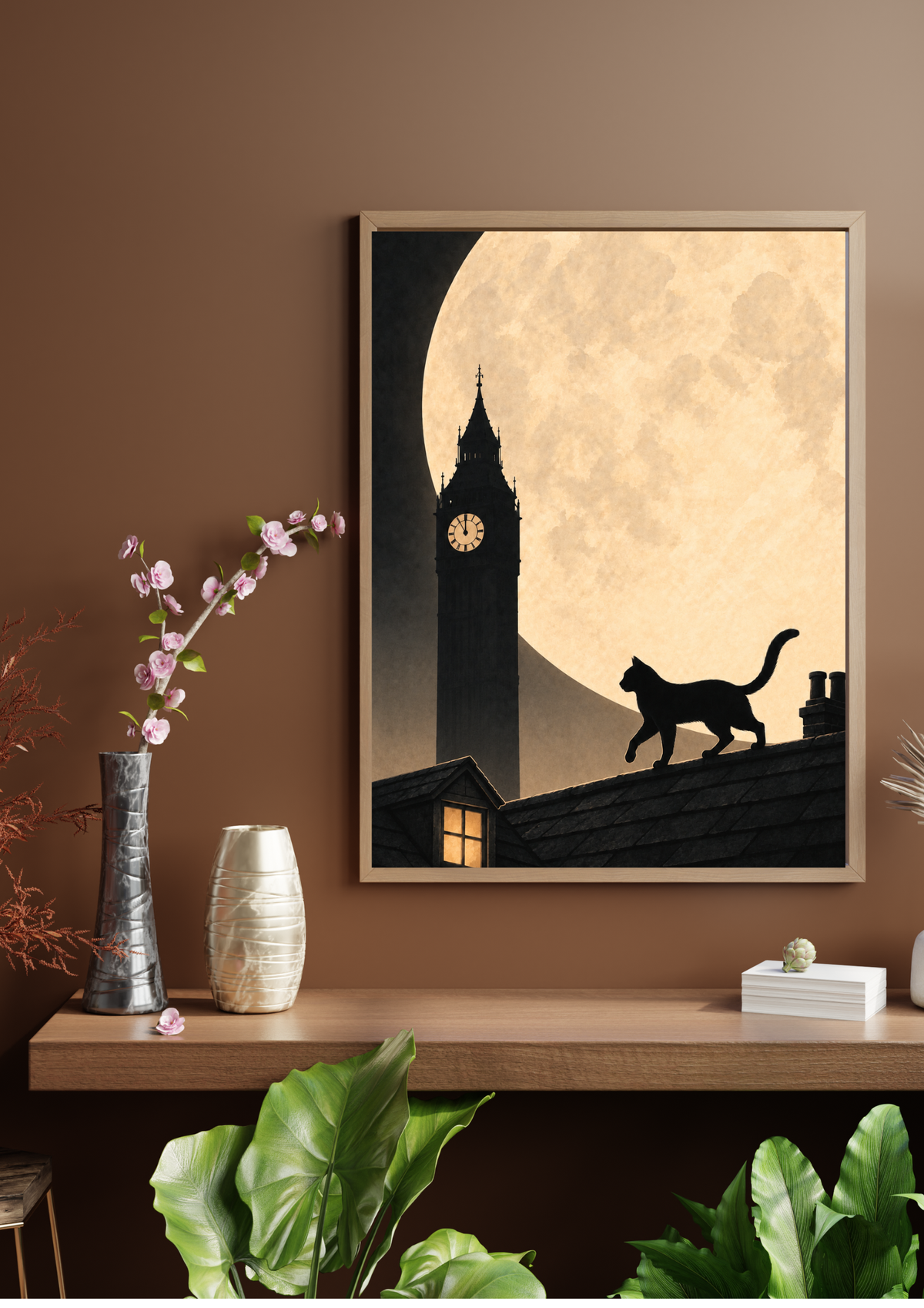 A framed vertical art print hanging on a brown wall above a wooden console table. The print shows a black silhouette of a cat walking across a roof in front of a large cream-colored full moon and a clock tower, with a small glowing window.
