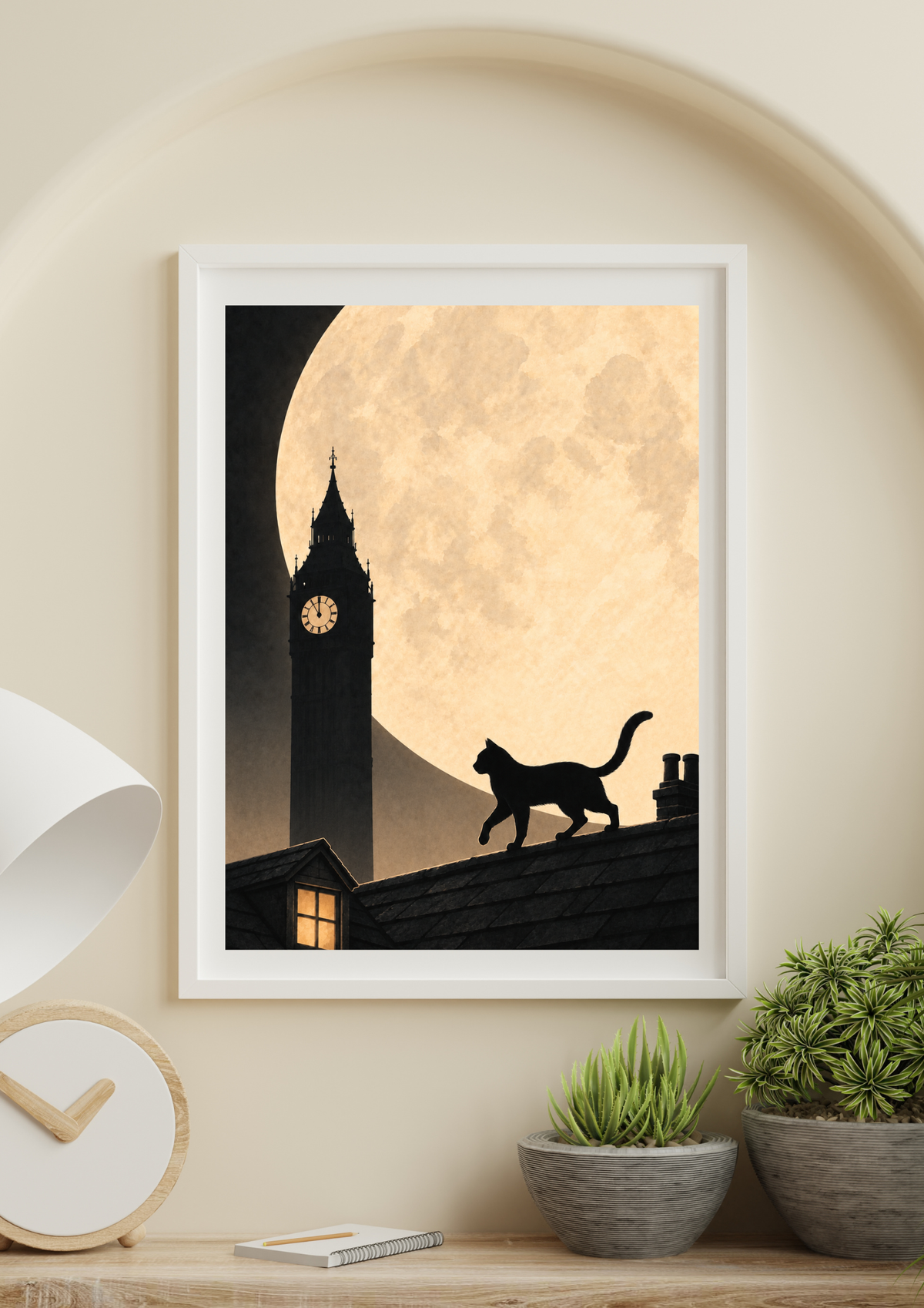A framed art print featuring a black cat silhouette walking along a roof against a large cream-colored moon and a dark clock tower, displayed on a wooden shelf in a styled room setting.