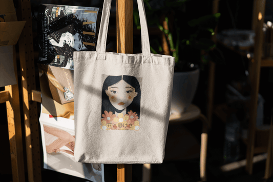 Canvas tote bag featuring a serene Japanese-style portrait of a woman surrounded by pastel flowers, hanging on a wooden rack in a softly lit room.