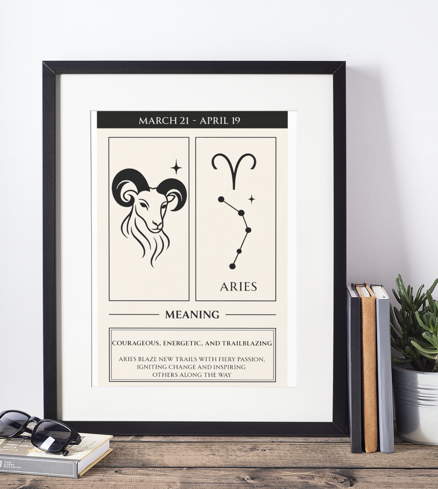 Mockup of a black-framed Aries zodiac poster standing on a rustic wood table against a white wall, flanked by stacked books, a small potted succulent, and a pair of sunglasses, featuring the Aries ram illustration, glyph, constellation, date range, and meaning text.