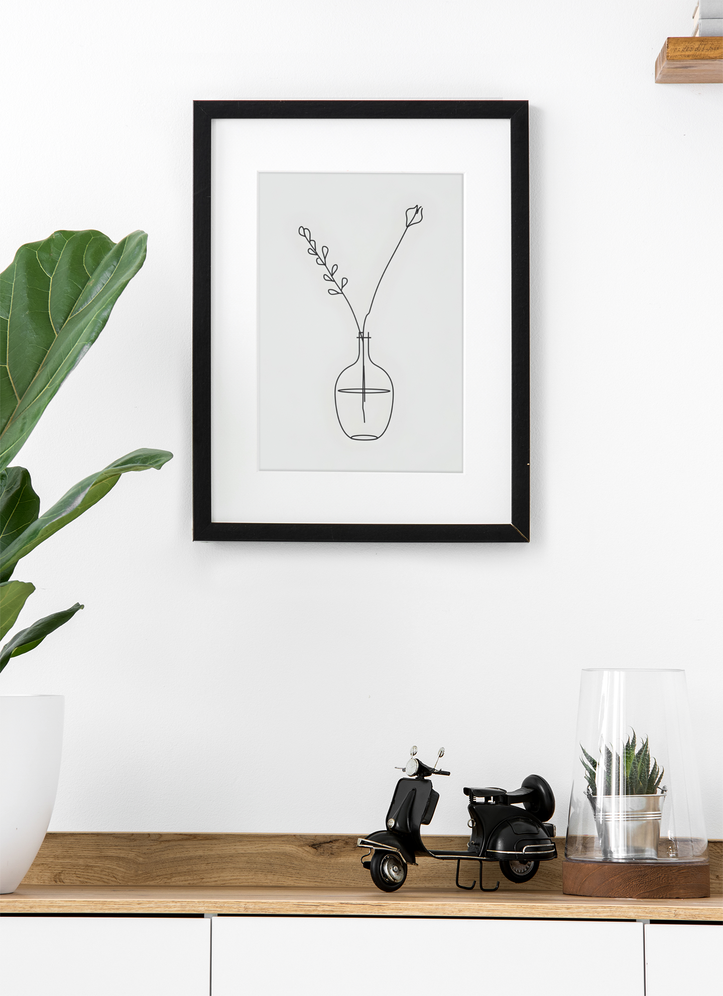 Framed minimalist line art of a vase with flowers on a white wall above a wooden shelf.
