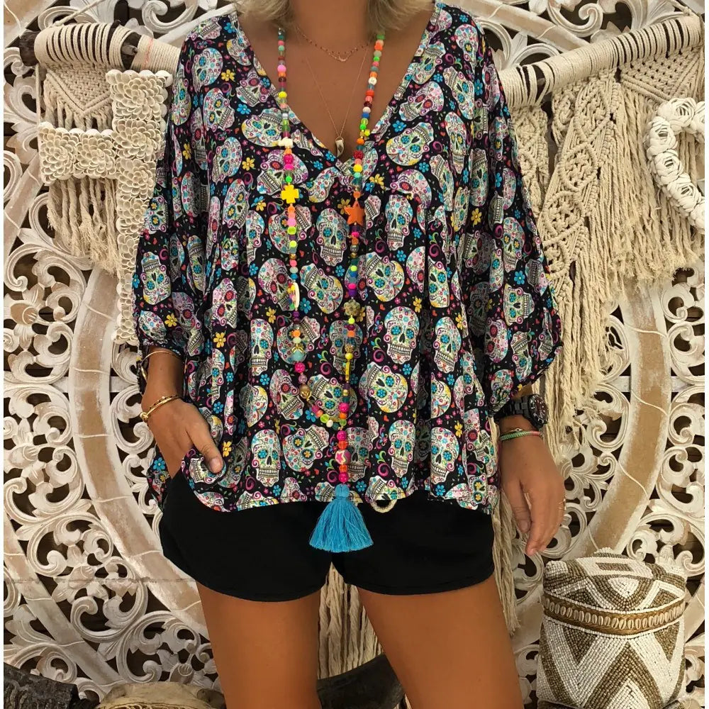 Person wearing a colorful skull patterned top with tassels in front of a decorative wall.