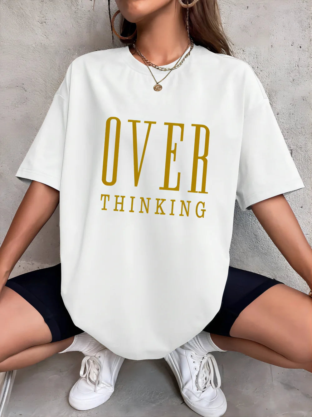 Person wearing a white t-shirt with 'OVER THINKING' in gold letters, sitting on the ground.