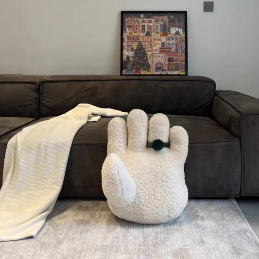 White fluffy hand-shaped pillow on a gray carpet with a brown sofa and framed picture in the background.
