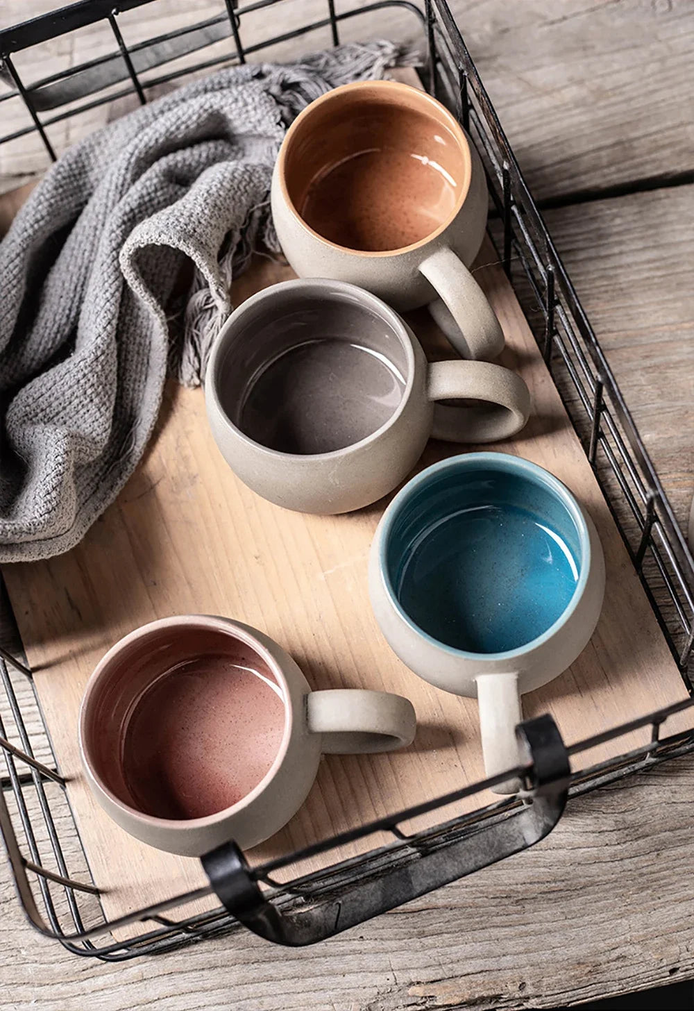 Set of ceramic mugs in various colors on a wooden surface with a wire basket.