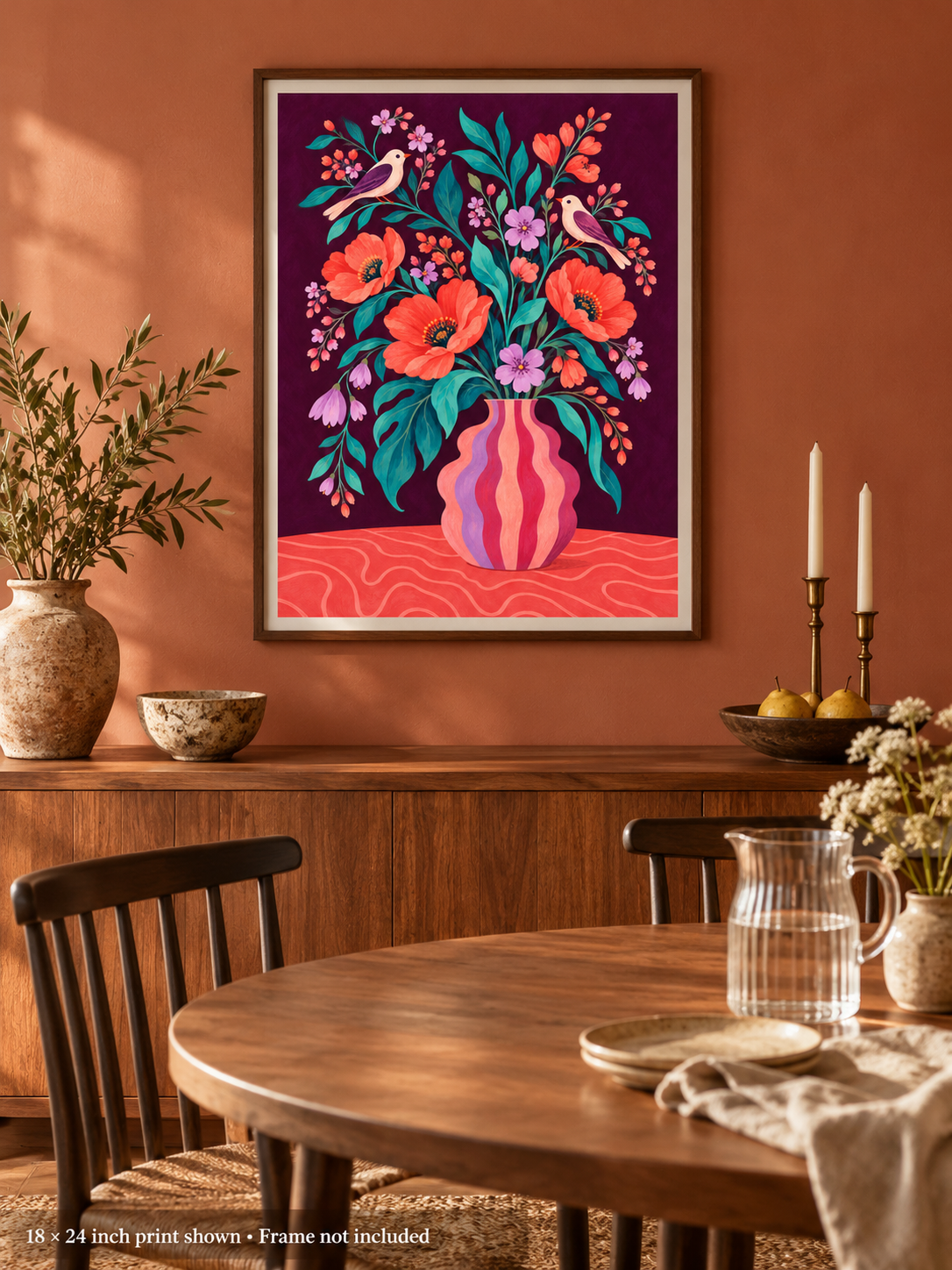 A framed art print featuring a colorful floral bouquet in a pink striped vase against a dark purple background, displayed on a terracotta wall above a wooden dining table in a styled room.
