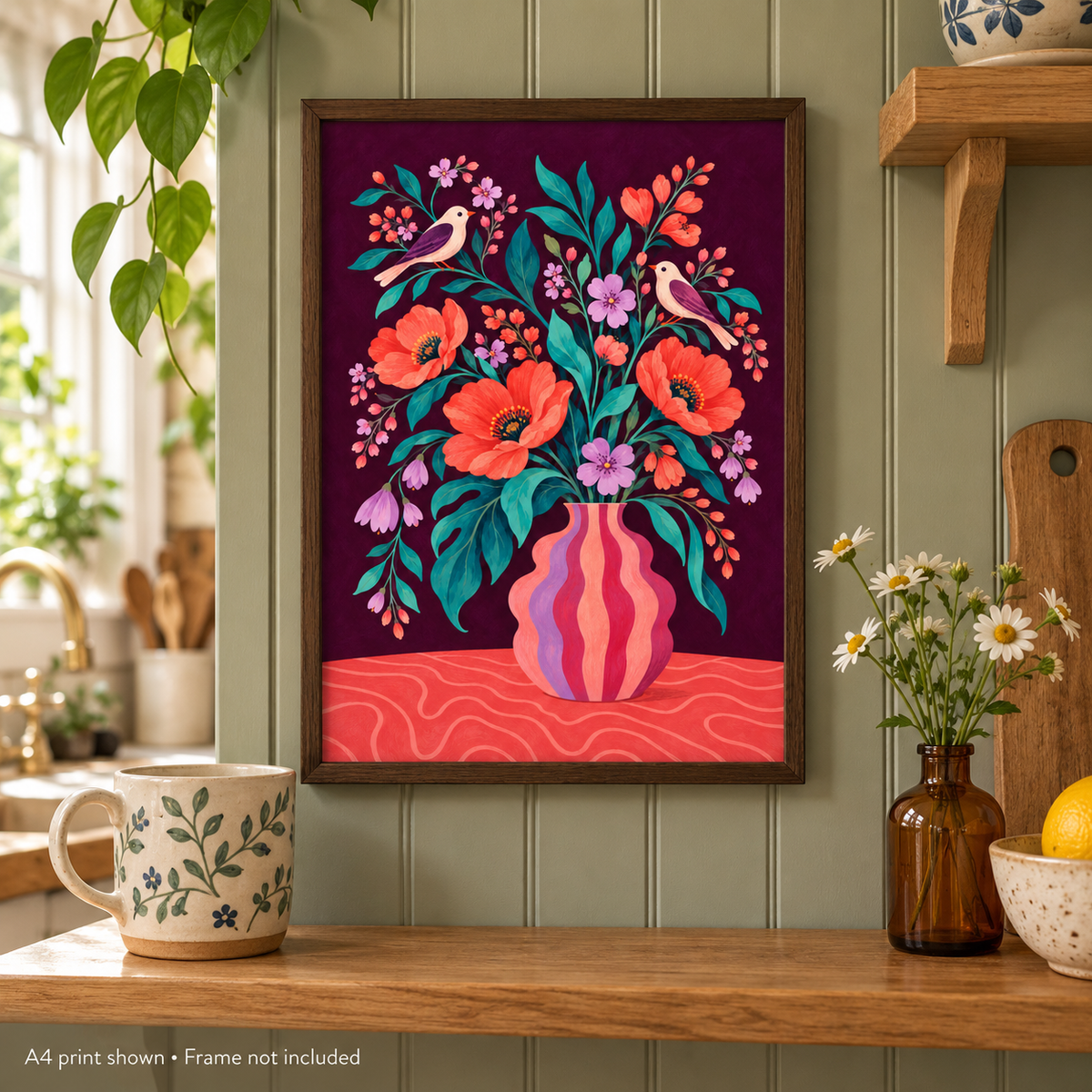 A framed art print featuring a colorful floral and bird illustration in a vase, displayed on a kitchen wall above a wooden shelf with a mug, flowers, and a cutting board.