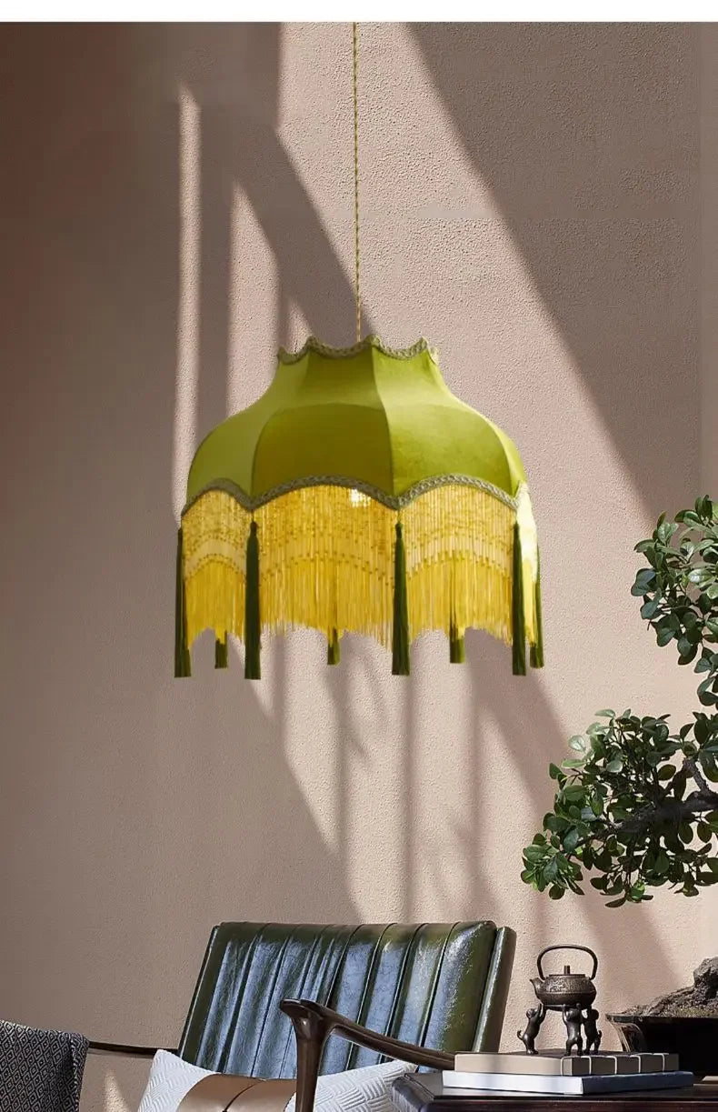 Green lampshade with yellow fringe hanging in a room with a plant and chair.
