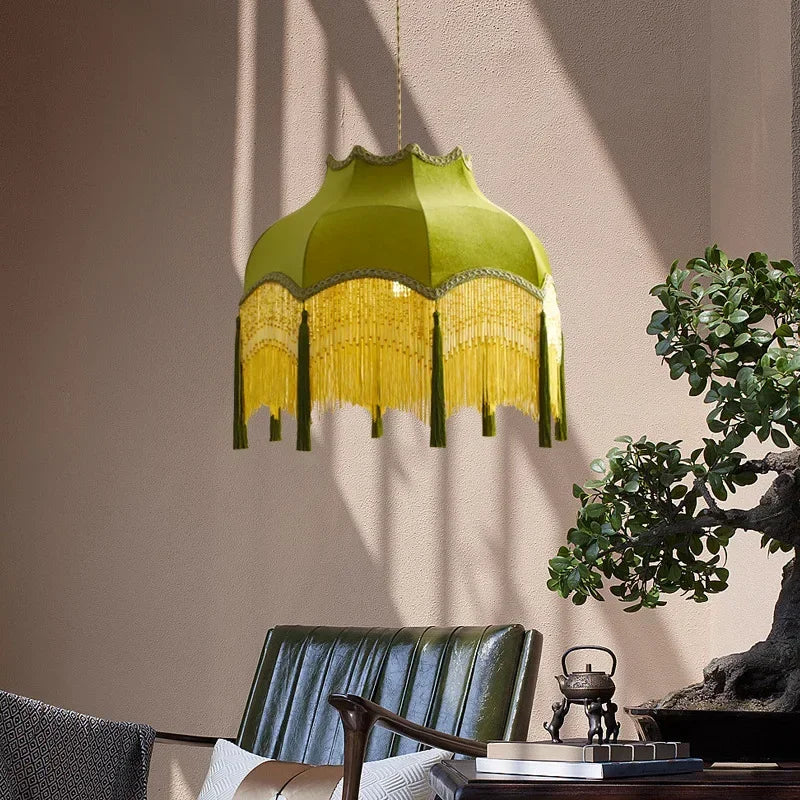 Green fringed lampshade hanging above a table with decorative items and a plant in the background.