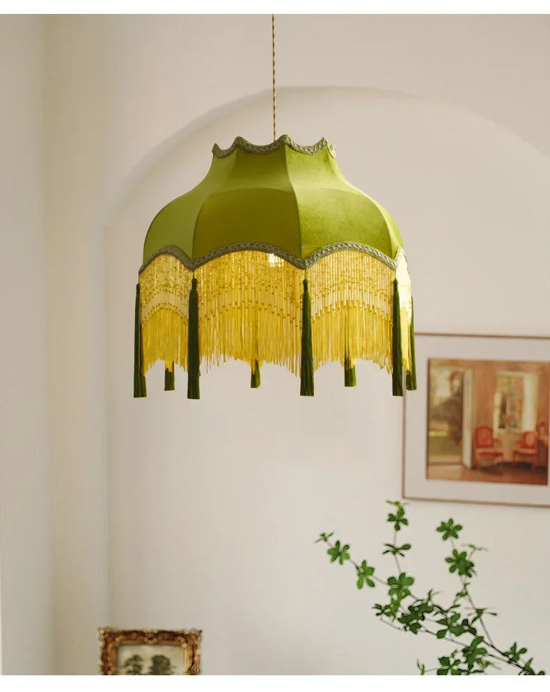 Green pendant lamp with fringe details hanging in a room with a plant and framed picture on the wall.