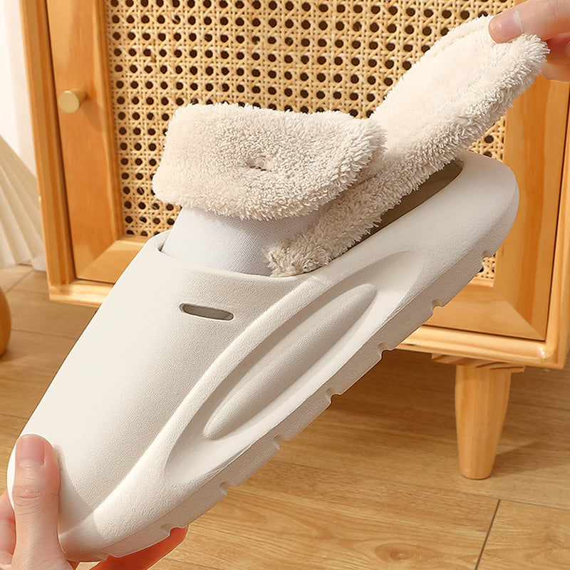 White slipper with fluffy white lining held by a hand on a wooden floor.