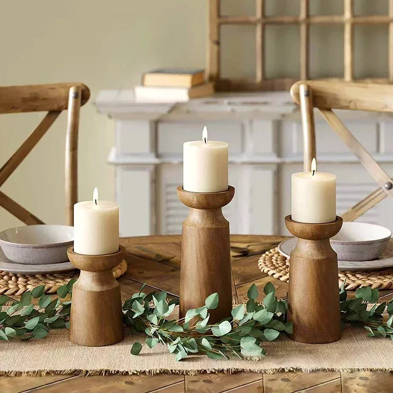 Three wooden candle holders with candles on a table with greenery and chairs in the background.
