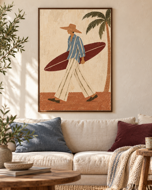 A framed wall art print featuring a retro-style illustration of a man carrying a surfboard, hung above a beige sofa with blue and burgundy pillows in a sunlit living room.