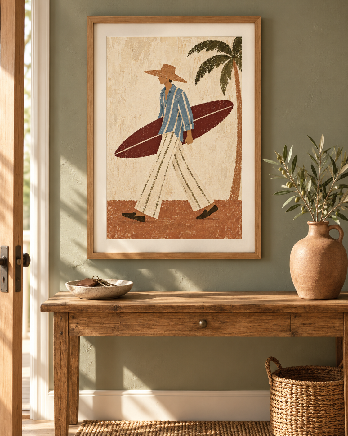 Framed wall art featuring a retro-style illustration of a surfer walking with a surfboard, hung on a sage green wall above a rustic wooden console table with a vase and basket.