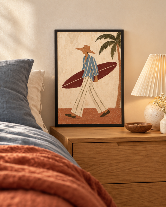 A framed art print featuring a retro-style illustration of a man carrying a surfboard under his arm, styled on a wooden nightstand next to a bed with blue and rust-colored bedding.