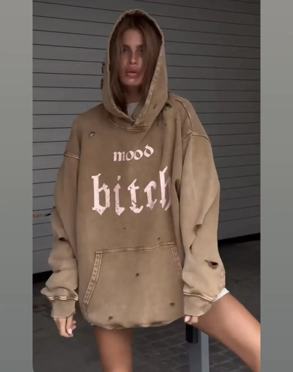 Person wearing a beige hoodie with text, standing against a gray wall.