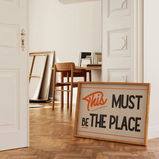 Framed sign with 'This Must Be The Place' text in a room with wooden floor and white walls.