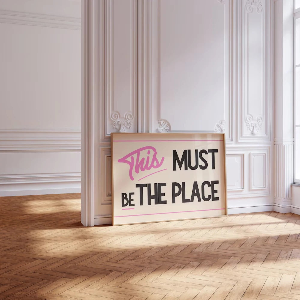 Framed quote 'This MUST BE THE PLACE' on a wall in an elegant room with wooden floor and white walls.