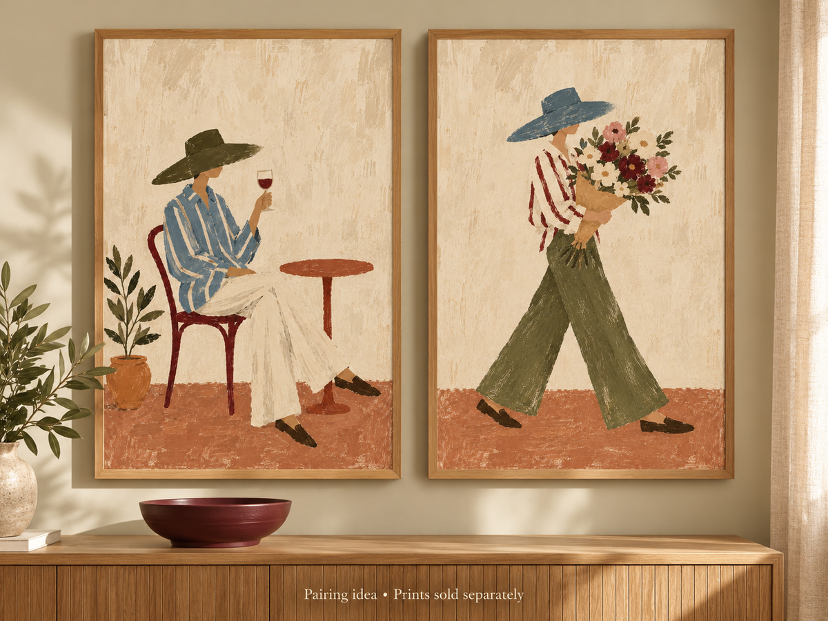 Two framed art prints displayed side-by-side on a beige wall above a wooden console table. The left print shows a seated person in a blue striped shirt and white pants holding a glass of red wine, while the right print shows a walking person in a red-and-white striped shirt and green pants carrying a bouquet of red, pink, and white flowers.