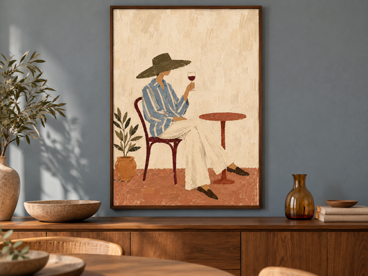 A framed art print depicting a woman in a blue striped shirt and white pants sitting at a table with a glass of red wine, hanging on a blue-gray wall above a wooden sideboard with decorative plants and vases.
